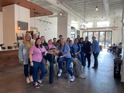 Группа улыбающихся людей в повседневной одежде собралась в светлом современном кафе с высокими потолками и большими окнами.