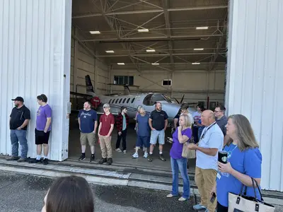 Группа людей стоит у открытой двери ангара, внутри которого частично виден небольшой самолет.