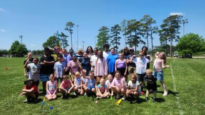 Группа улыбающихся детей и взрослых позирует в солнечный день в парке, на фоне зеленой травы и высоких деревьев, передавая радость и товарищество.