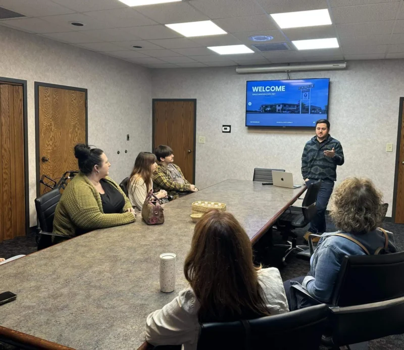 Мужчина стоит в конце стола для совещаний и разговаривает с пятью сидящими людьми.