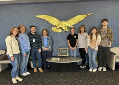 Группа из восьми человек стоит, улыбаясь, перед большим золотым орлом на голубой стене.