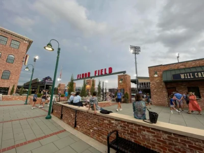 Сцена на открытом воздухе на поле Fluor Field, где люди собираются у кирпичных строений и фонарных столбов под пасмурным небом.